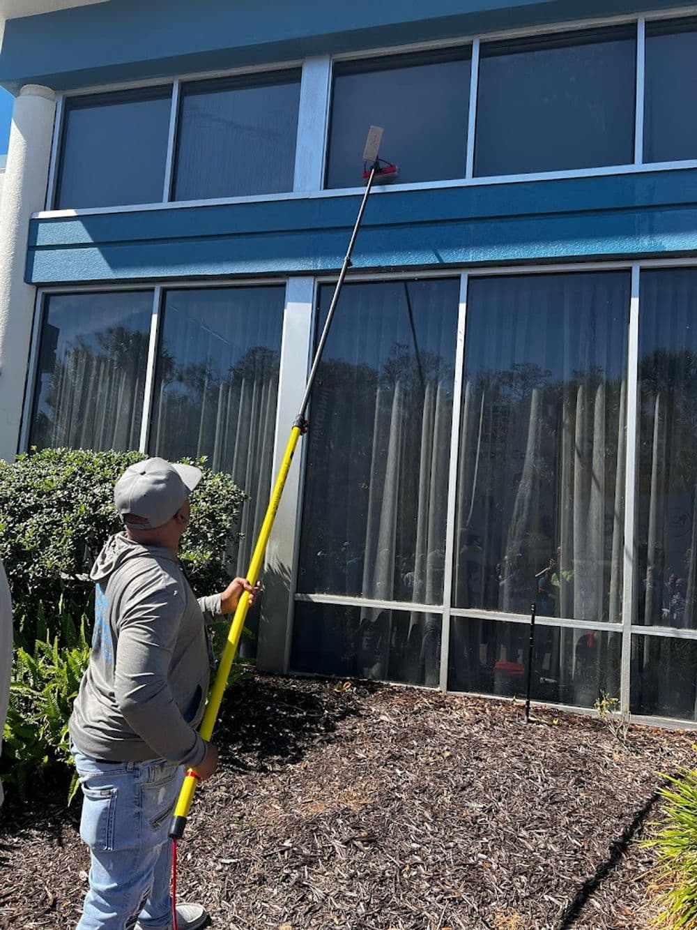 Person cleaning a high window using a long reach pole and squeegee outside a commercial building.