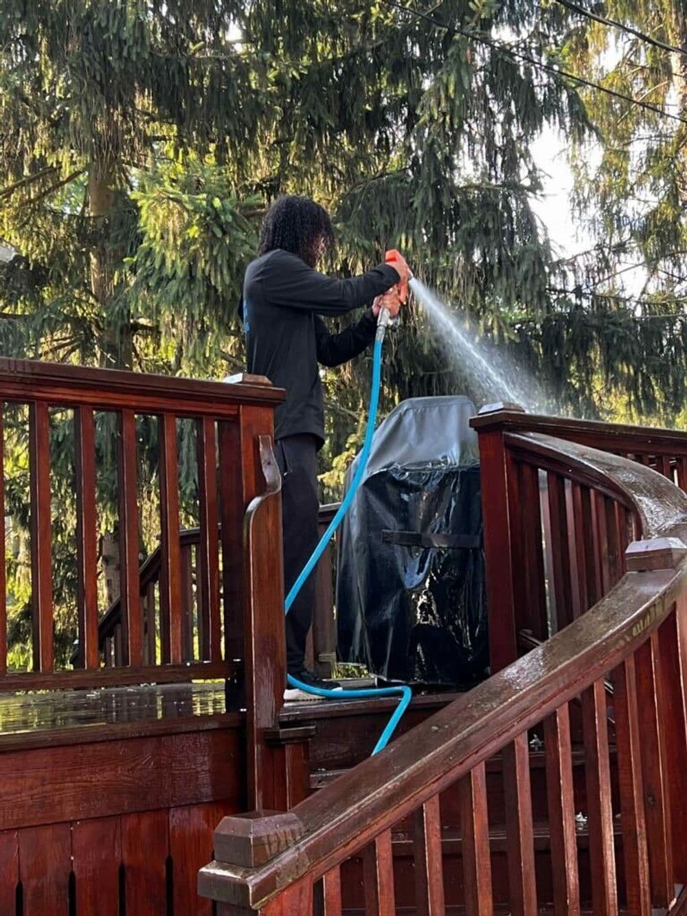 Person cleaning a barbecue grill with a hose on a wooden deck surrounded by trees.